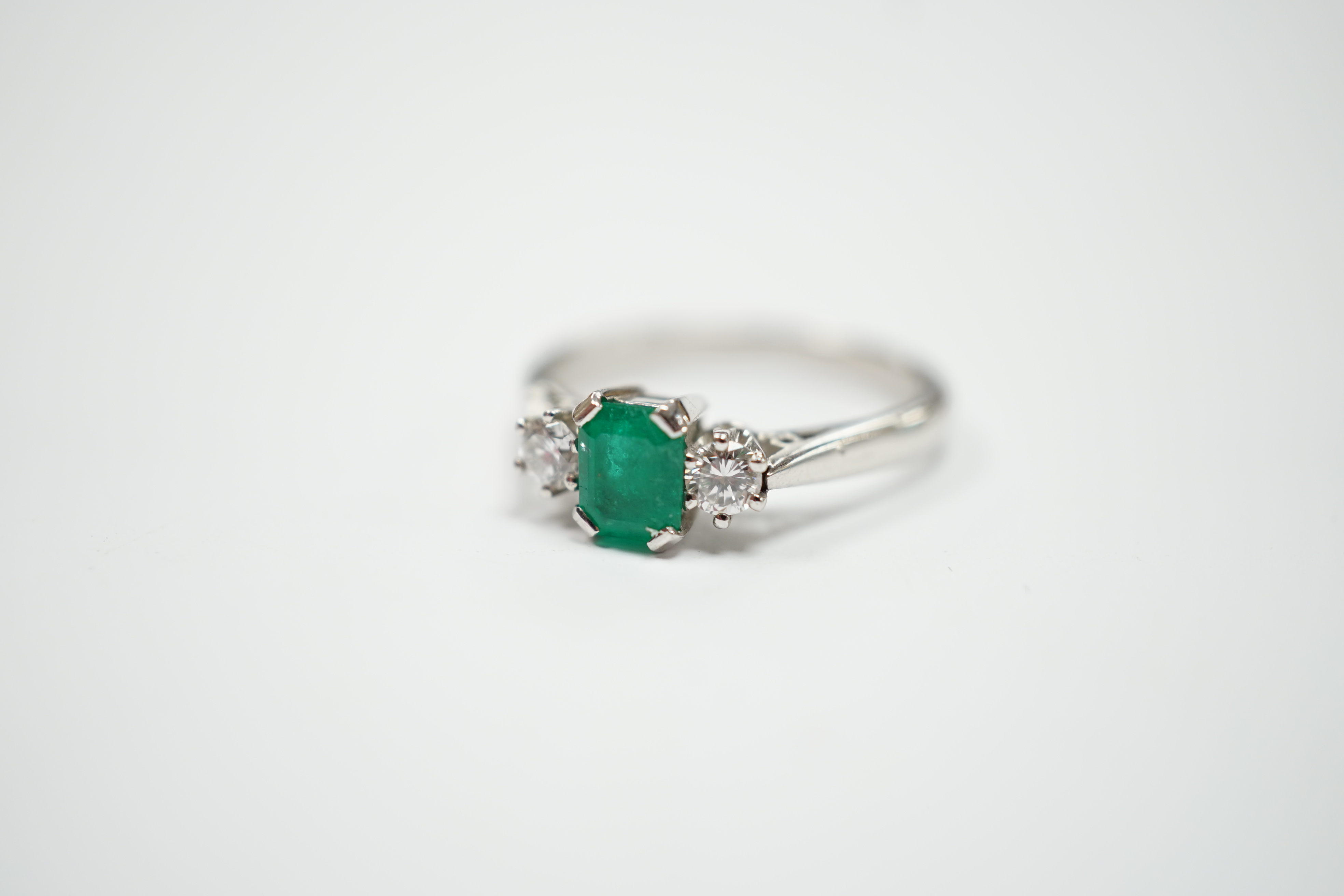A modern 18ct white metal, single stone emerald and two stone diamond set ring, size N, gross weight 4 gram.